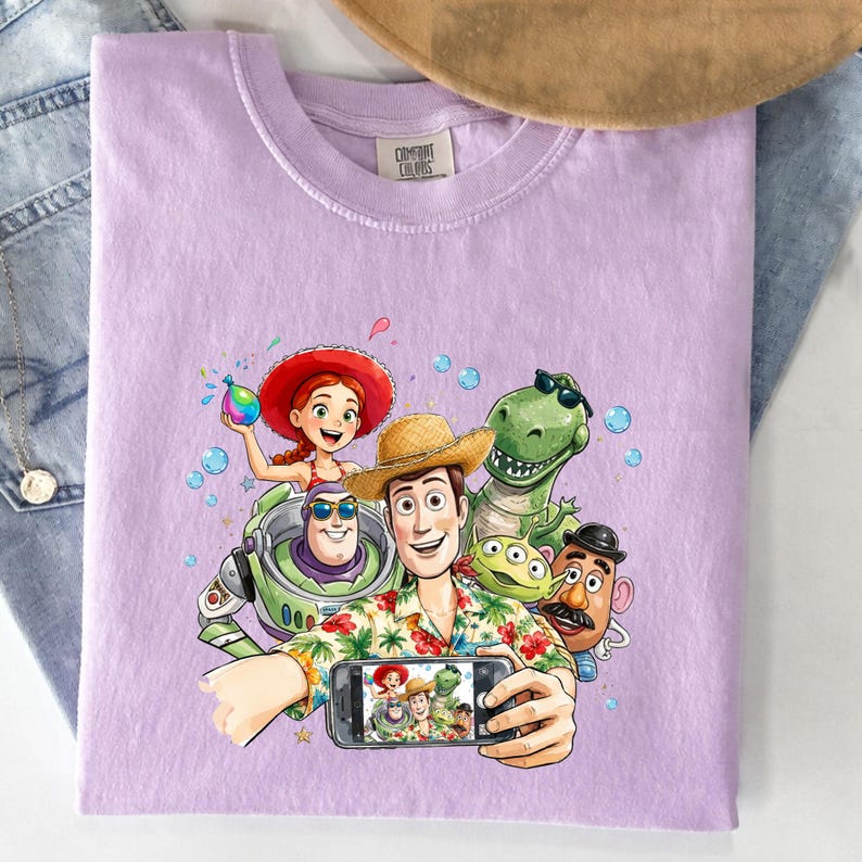 May include: A light purple t-shirt featuring a colorful graphic of characters from the animated movie Toy Story. The design includes Woody, Buzz Lightyear, Rex, Jessie, and others taking a selfie. The shirt is folded.