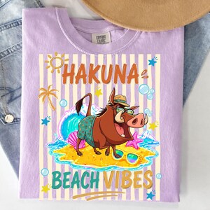 May include: Lilac Comfort Colors t-shirt with a graphic featuring a cartoon warthog wearing sunglasses and a hat, with the words "HAKUNA BEACH VIBES". The design includes a sun, palm tree, waves, and starfish.