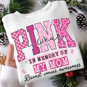 K&ouml;nnte beinhalten: Wei&szlig;es Sweatshirt mit dem Text "PINK I wear" in rosa Blockbuchstaben mit verschiedenen Mustern. Die Worte "IN MEMORY OF MY MOM" und "Breast cancer awareness" sind ebenfalls auf dem Shirt aufgedruckt. Rosa Schleifen und Blumen sind enthalten.