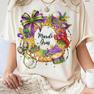 May include: Cream-colored t-shirt featuring a vibrant Mardi Gras wreath design. The wreath includes a purple and green bow, a mask, a crawfish, a drum, and the words "Mardi Gras". The design is embellished with gold and purple beads and accents.