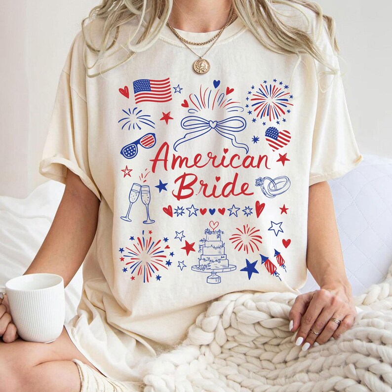 May include: Cream-colored t-shirt with a patriotic design featuring an American flag, fireworks, and the text "American Bride" in red and blue. The shirt has a relaxed fit and short sleeves.