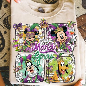 May include: Beige t-shirt featuring a colorful Mardi Gras design. The design includes Mickey Mouse, Minnie Mouse, Goofy, and Pluto in festive attire, with the words "Mardi Gras" prominently displayed.