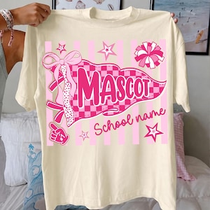 May include: A cream-colored t-shirt featuring a pink graphic design with a checkered pennant flag, the word MASCOT, and the text School name. The design includes a large bow, a pom-pom, stars, and a foam finger on a background of vertical stripes.