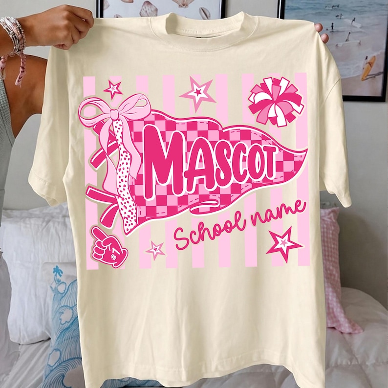 May include: A cream-colored t-shirt featuring a pink graphic design with a checkered pennant flag, the word MASCOT, and the text School name. The design includes a large bow, a pom-pom, stars, and a foam finger on a background of vertical stripes.