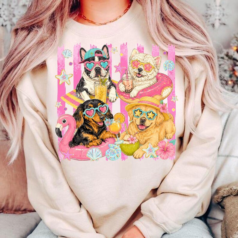 May include: A beige sweatshirt featuring a graphic of four dogs enjoying summer. The dogs wear sunglasses and are surrounded by beach-themed elements like a flamingo float, drinks, and flowers. The background has pink and white stripes.
