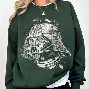 May include: Dark green sweatshirt featuring a detailed graphic of Darth Vader's helmet and space battle elements. The design is in white, creating a striking contrast against the dark green fabric. The sweatshirt has long sleeves and a crew neck.