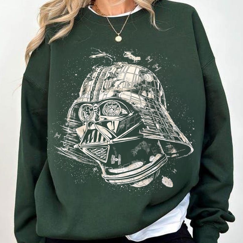 May include: Dark green sweatshirt featuring a detailed graphic of Darth Vader's helmet and space battle elements. The design is in white, creating a striking contrast against the dark green fabric. The sweatshirt has long sleeves and a crew neck.
