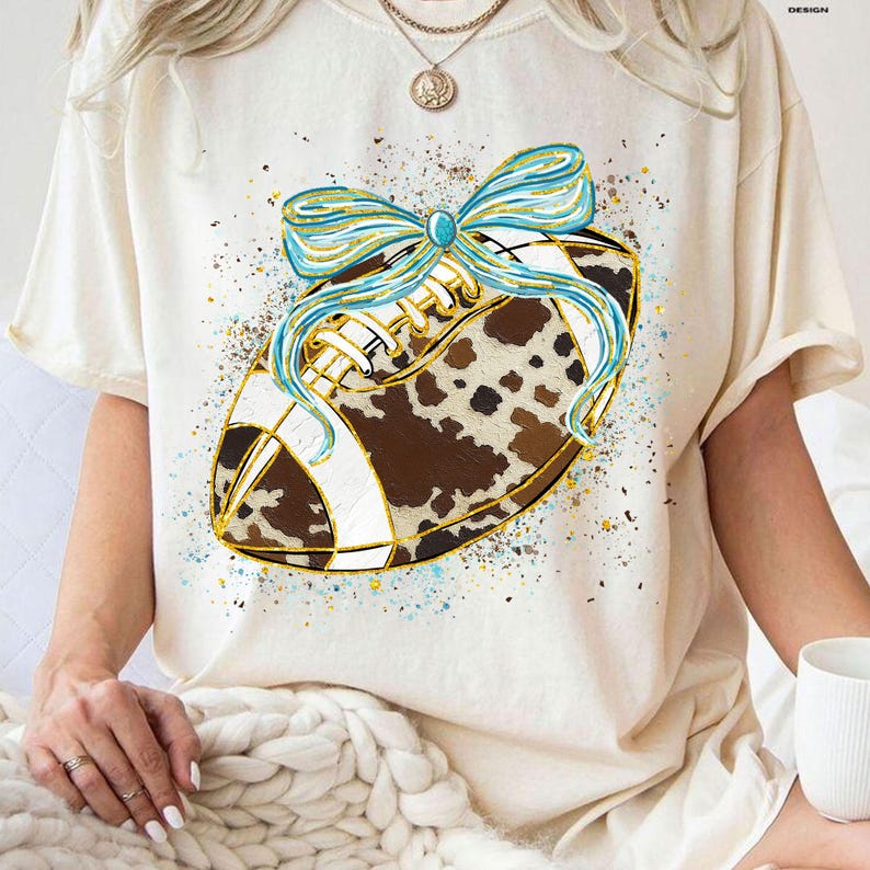 May include: Cream-colored t-shirt featuring a football design. The football has a brown and white cowhide pattern, accented with gold trim and a light blue bow. The design is embellished with blue and gold paint splatters.