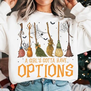 May include: White sweatshirt featuring a design of six different witch brooms, spiderwebs, and bats. The text "A Girl's Gotta Have Options" is printed in orange. A Halloween-themed graphic.