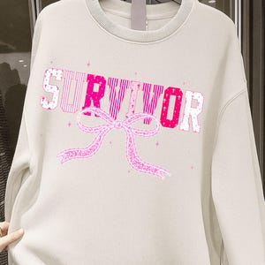 May include: Cream-colored sweatshirt with the word "SURVIVOR" in pink, with a pink ribbon graphic. The letters have different patterns, and small pink stars are scattered around the design. The sweatshirt has a crew neck.