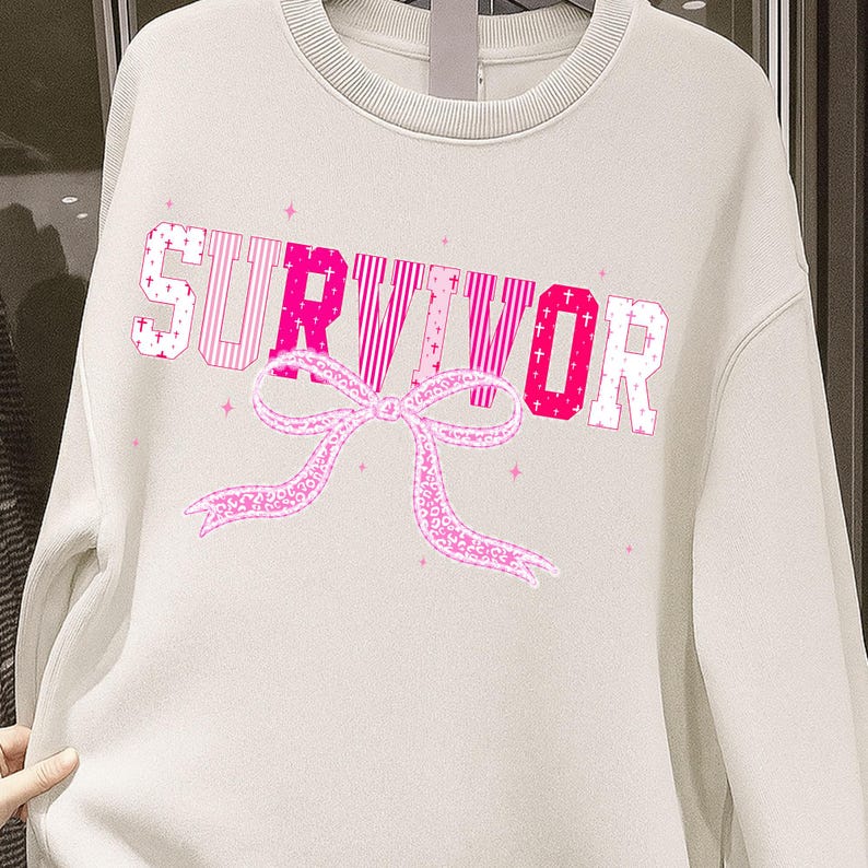 May include: Cream-colored sweatshirt with the word "SURVIVOR" in pink, with a pink ribbon graphic. The letters have different patterns, and small pink stars are scattered around the design. The sweatshirt has a crew neck.