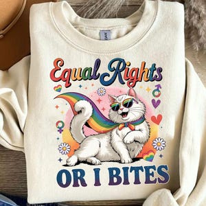 May include: Cream-colored sweatshirt with a graphic of a white cat wearing sunglasses and a rainbow cape. The text "Equal Rights OR I BITES" is displayed above and below the cat illustration, with various pride symbols.