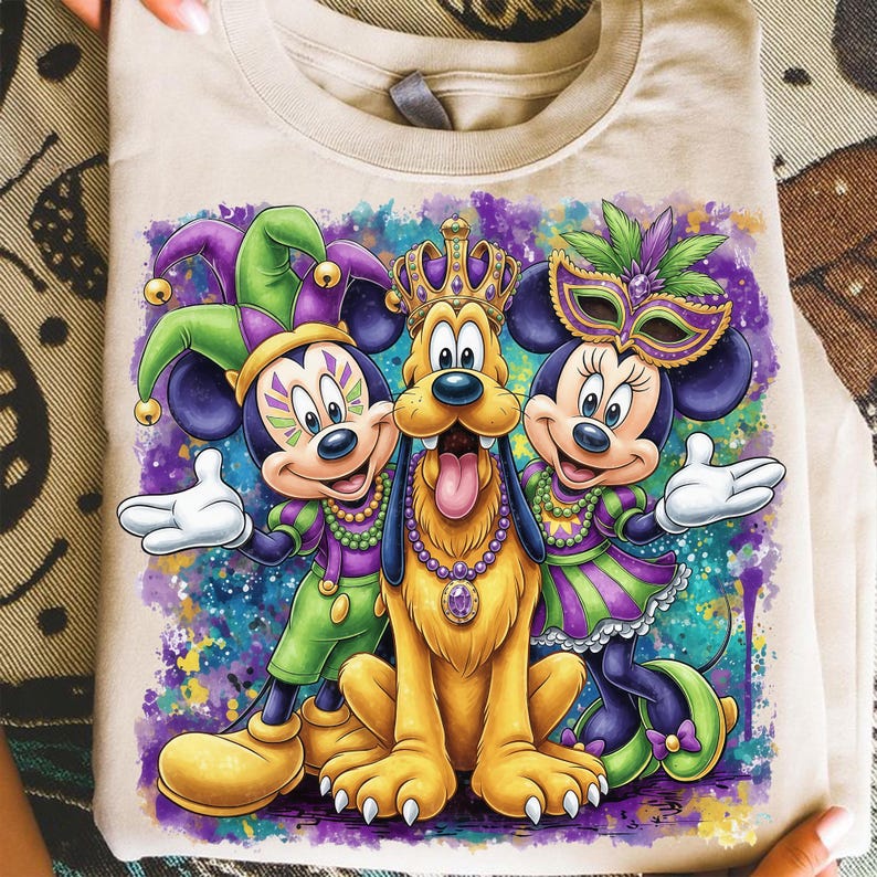 May include: A beige t-shirt featuring a colorful graphic of Mickey Mouse, Pluto, and Minnie Mouse in Mardi Gras attire. Mickey and Minnie wear masks and jester hats, while Pluto sports a crown. The design has a vibrant, watercolor-like background.