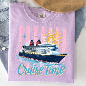 May include: A lilac t-shirt featuring a cruise ship design. The graphic includes a blue and white cruise ship, a yellow sun, pink stripes, and the words "Cruise Time" in blue and teal. The shirt is made of a soft material.