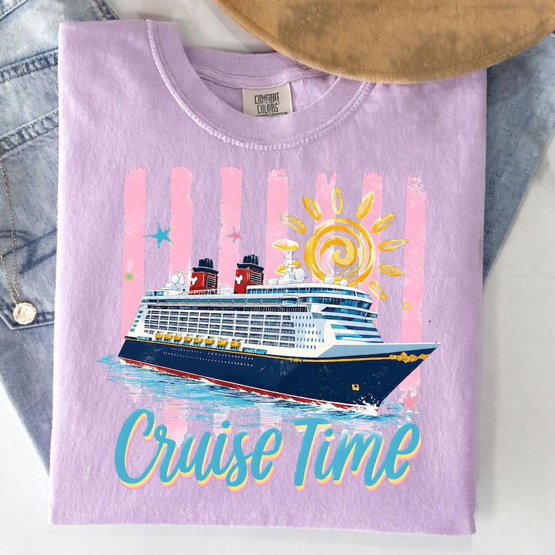 May include: A lilac t-shirt featuring a cruise ship design. The graphic includes a blue and white cruise ship, a yellow sun, pink stripes, and the words "Cruise Time" in blue and teal. The shirt is made of a soft material.