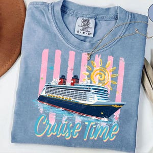 May include: A pale blue t-shirt featuring a cruise ship design. The graphic includes a cruise ship, a yellow sun, pink stripes, and the words "Cruise Time" in a colourful font. The shirt has a crew neck and short sleeves.