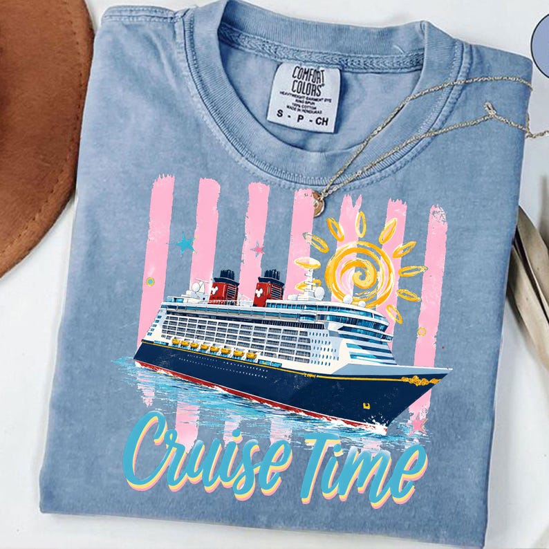 May include: A pale blue t-shirt featuring a cruise ship design. The graphic includes a cruise ship, a yellow sun, pink stripes, and the words "Cruise Time" in a colourful font. The shirt has a crew neck and short sleeves.