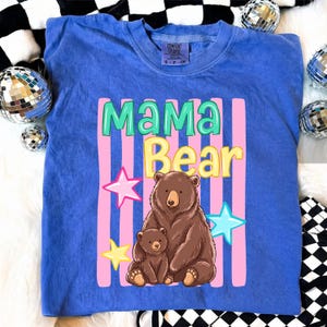 Pu&ograve; includere: Una maglietta blu con la scritta "MAMA Bear" in un design colorato. La grafica include un orso marrone e un cucciolo di orso pi&ugrave; piccolo, con strisce rosa e stelle. La maglietta &egrave; del marchio Comfort Colors.