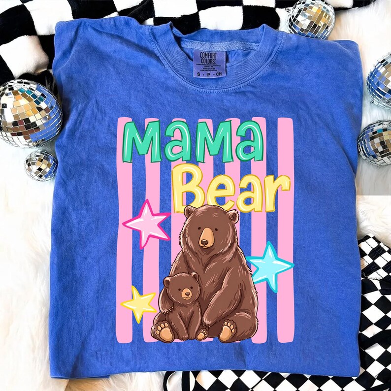 Pu&ograve; includere: Una maglietta blu con la scritta "MAMA Bear" in un design colorato. La grafica include un orso marrone e un cucciolo di orso pi&ugrave; piccolo, con strisce rosa e stelle. La maglietta &egrave; del marchio Comfort Colors.