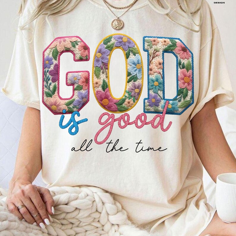 Op de afbeelding: Cr&egrave;mekleurig T-shirt met de tekst "GOD is good all the time" in een bloemenontwerp. De letters zijn gevuld met kleurrijke bloemen, de tekst is een mix van script- en blokletters.