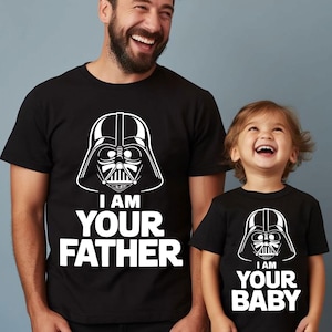 May include: Matching black t-shirts with white graphics. One shirt features a Darth Vader helmet design with the text "I AM YOUR FATHER." The other shirt has the same design with the text "I AM YOUR BABY."