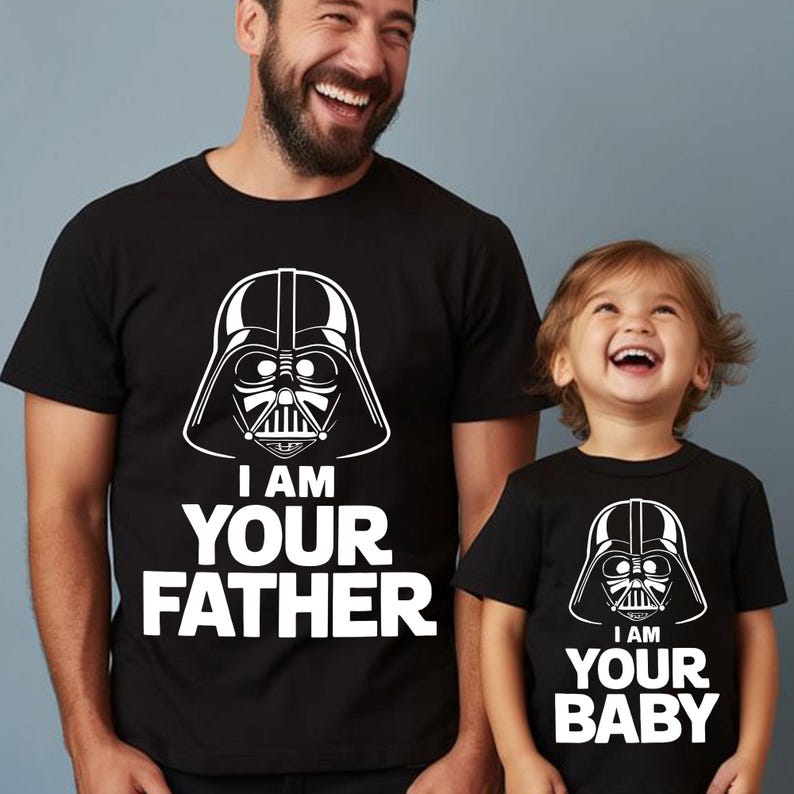 May include: Matching black t-shirts with white graphics. One shirt features a Darth Vader helmet design with the text "I AM YOUR FATHER." The other shirt has the same design with the text "I AM YOUR BABY."