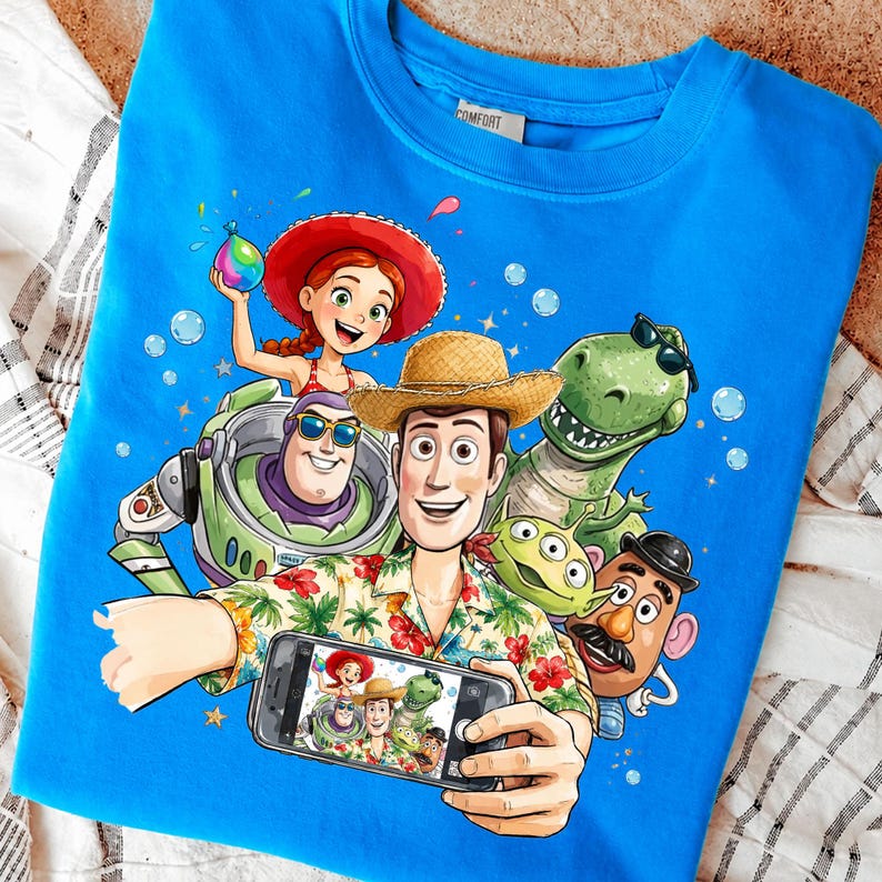 Może przedstawiać: Niebieska bluza z kolorową grafiką postaci z kresk&oacute;wek robiących sobie selfie. Projekt zawiera kowboja, kosmicznego strażnika, dinozaura i inne animowane postacie. Kowboj trzyma telefon. Bluza ma metkę z napisem "Comfort".