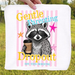 Op de afbeelding: Witte vierkante handdoek met een wasbeer met een zonnebril en een koffiekopje. De tekst "Gentle Parenting Dropout" staat in geel en blauw, met roze verticale strepen en sterren.
