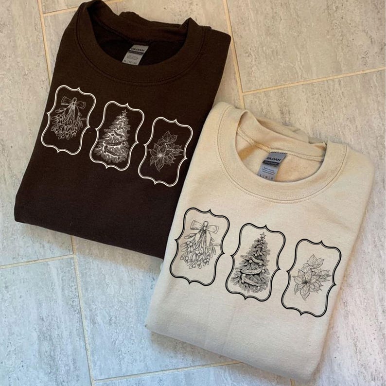 May include: Two sweatshirts, one brown and one beige, each featuring a decorative design. The design includes a mistletoe sprig, a Christmas tree, and a poinsettia, all within ornate frames. The sweatshirts are folded.