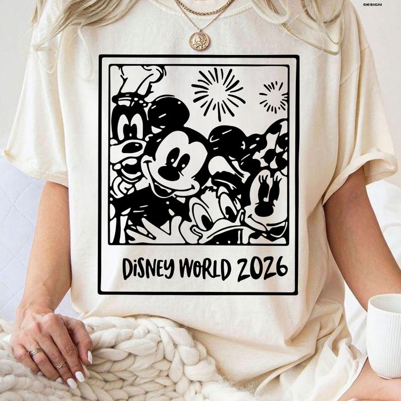 Op de afbeelding: Cr&egrave;mekleurig T-shirt met een zwart-witte afbeelding van Disney-personages, waaronder Mickey Mouse, Minnie Mouse, Donald Duck en Goofy. De tekst "DISNEY WORLD 2026" staat onder de afbeelding.