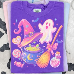Puede incluir: Una camiseta morada con un diseño caprichoso de Halloween. El gráfico incluye un sombrero de bruja, un caldero, un fantasma, una escoba y una botella de poción con el texto "Spooky Cutie". El diseño también presenta piruletas, estrellas y murciélagos.