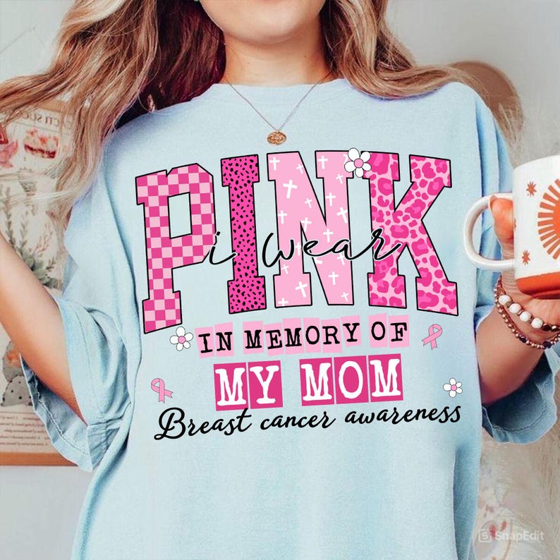 K&ouml;nnte beinhalten: Hellblaues T-Shirt mit den Worten "PINK I WEAR" in rosa, karierten und leopardenmusternden Buchstaben. Darunter steht der Text "IN MEMORY OF MY MOM", darunter "Breast cancer awareness". Eine Tasse wird in der rechten Hand gehalten.