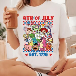 May include: White t-shirt with a graphic of Toy Story characters wearing red, white, and blue patriotic hats. The text "4th of July" and "Est. 1776" is printed on the shirt.