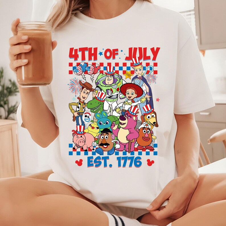 May include: White t-shirt with a graphic of Toy Story characters wearing red, white, and blue patriotic hats. The text "4th of July" and "Est. 1776" is printed on the shirt.