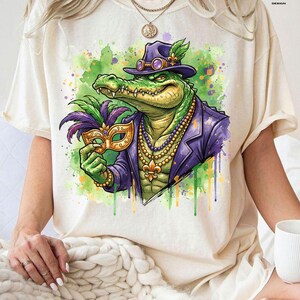 May include: Cream-colored t-shirt featuring a vibrant illustration of an alligator in a purple suit and hat, holding a gold mask. The alligator wears a beaded necklace and is set against a watercolor-style background with green and purple splashes.