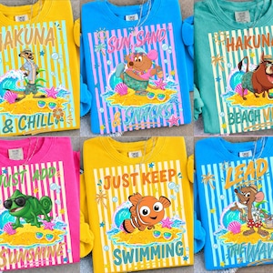 May include: A collection of colorful sweatshirts featuring cartoon characters and beach-themed phrases. Designs include "Hakuna & Chill," "Sun, Sand, Snacks," and "Just Keep Swimming." Each shirt has a unique character illustration and striped background.