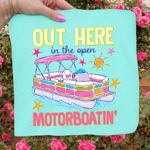 May include: A turquoise graphic featuring a colorful pontoon boat with the text "OUT HERE in the open MOTORBOATING'". The boat has a pink canopy and is surrounded by stars and a sun illustration. The design is held by a hand.