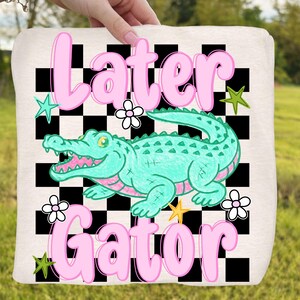 May include: A light-colored tote bag featuring a cartoon alligator in turquoise and pink, with the words "Later Gator" in pink bubble letters. The background has a black and white checkerboard pattern, with star and flower accents.