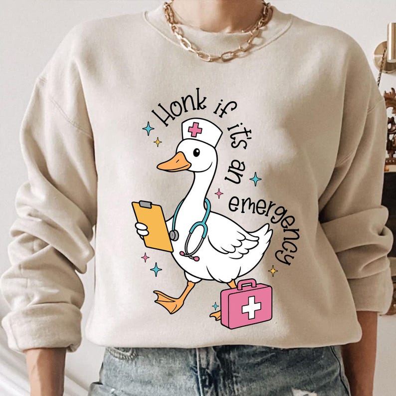 Puede incluir: Una sudadera beige con un dibujo animado de un ganso con un gorro de enfermera y sosteniendo un portapapeles y un estetoscopio. El texto "Honk if it's an emergency" est&aacute; impreso en la sudadera.