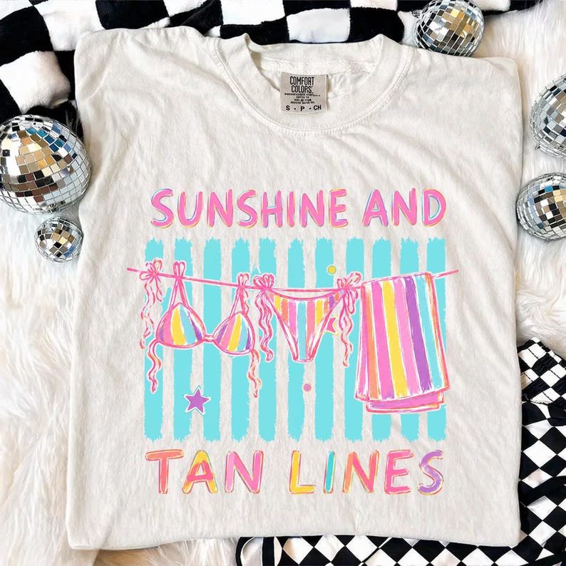 Peut inclure: T-shirt cr&egrave;me avec l'inscription "SUNSHINE AND TAN LINES" en rose et jaune. Le motif repr&eacute;sente un bikini et une serviette suspendus &agrave; une corde &agrave; linge avec des rayures verticales bleues. Le t-shirt a une &eacute;tiquette Comfort Colors.