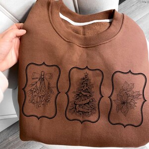 May include: A brown sweatshirt featuring three black framed illustrations: mistletoe, a Christmas tree, and poinsettia. The sweatshirt has a ribbed neckline and cuffs, and a white band inside the collar.