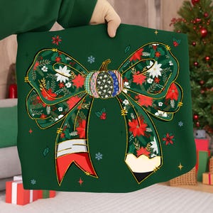 May include: A green fabric with a large bow design featuring a pumpkin center and floral patterns. The bow has red, white, and green floral accents, with a pencil design on the bottom. The design is held by a person.