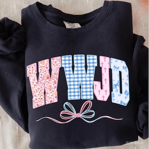 May include: A navy blue sweatshirt with the letters "WWJD" in a floral, gingham, and bow pattern. The letters are in pink, blue, and white. A pink and blue bow design is at the bottom.