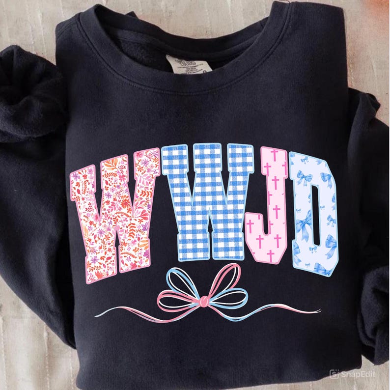 May include: A navy blue sweatshirt with the letters "WWJD" in a floral, gingham, and bow pattern. The letters are in pink, blue, and white. A pink and blue bow design is at the bottom.
