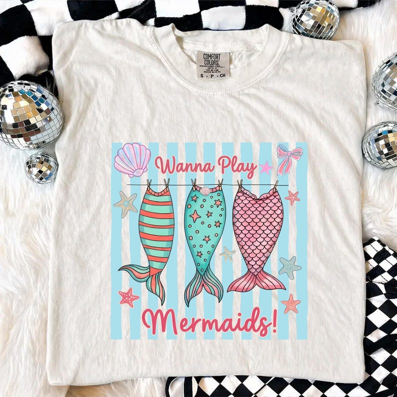 May include: Cream-colored t-shirt featuring a graphic of three mermaid tails hanging on a clothesline. The tails are striped, star-studded, and scaled in shades of teal, pink, and coral. The text "Wanna Play Mermaids!" is printed above and below the tails.