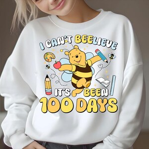 May include: White sweatshirt with a colorful graphic featuring a cartoon bear dressed as a bee, holding an apple. The text reads "I CAN'T BEELEIVE IT'S BEEN 100 DAYS" with school-themed illustrations.