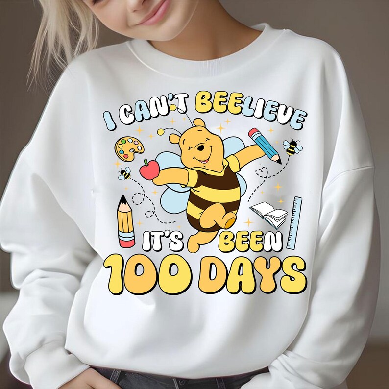 May include: White sweatshirt with a colorful graphic featuring a cartoon bear dressed as a bee, holding an apple. The text reads "I CAN'T BEELEIVE IT'S BEEN 100 DAYS" with school-themed illustrations.
