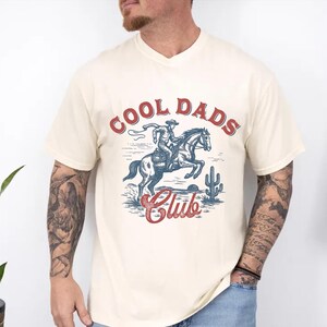 May include: Cream-colored V-neck t-shirt with a graphic of a cowboy riding a horse, a cactus, and the words "COOL DADS Club" in red and blue. The design has a vintage, Western aesthetic.