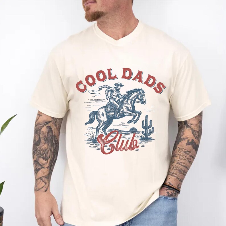May include: Cream-colored V-neck t-shirt with a graphic of a cowboy riding a horse, a cactus, and the words "COOL DADS Club" in red and blue. The design has a vintage, Western aesthetic.