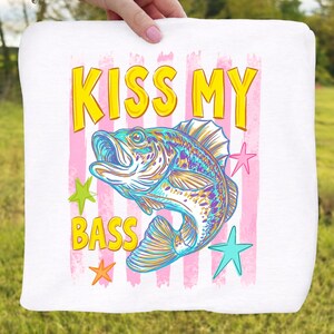 May include: White square fabric with the text "KISS MY BASS" in yellow and a colorful illustration of a fish. Pink vertical stripes and star shapes surround the fish. The fish is blue, yellow, and purple.
